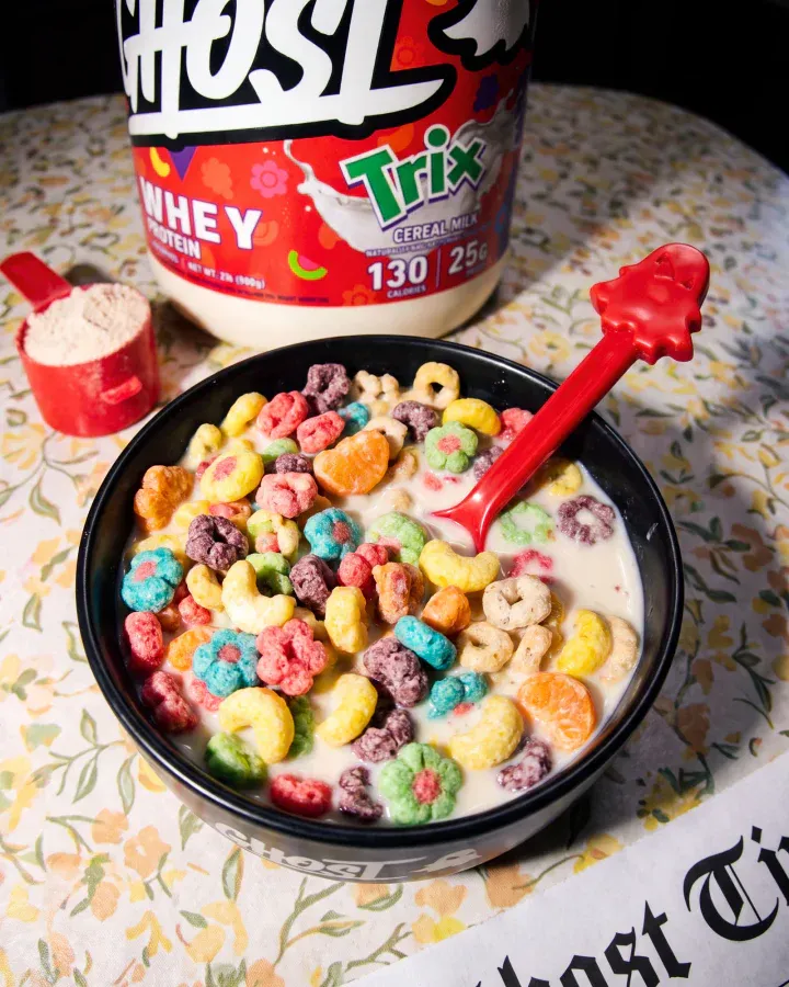 Ghost x Trix whey tub sits above a black bowl of colorful cereal with a red spoon, powder scoop and newspaper texture.