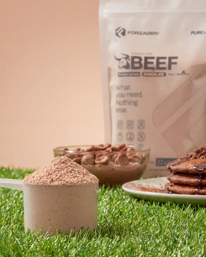 Scoop of Pure Beef Chocolate protein powder with the bag, pancakes and chocolate on a grass set.