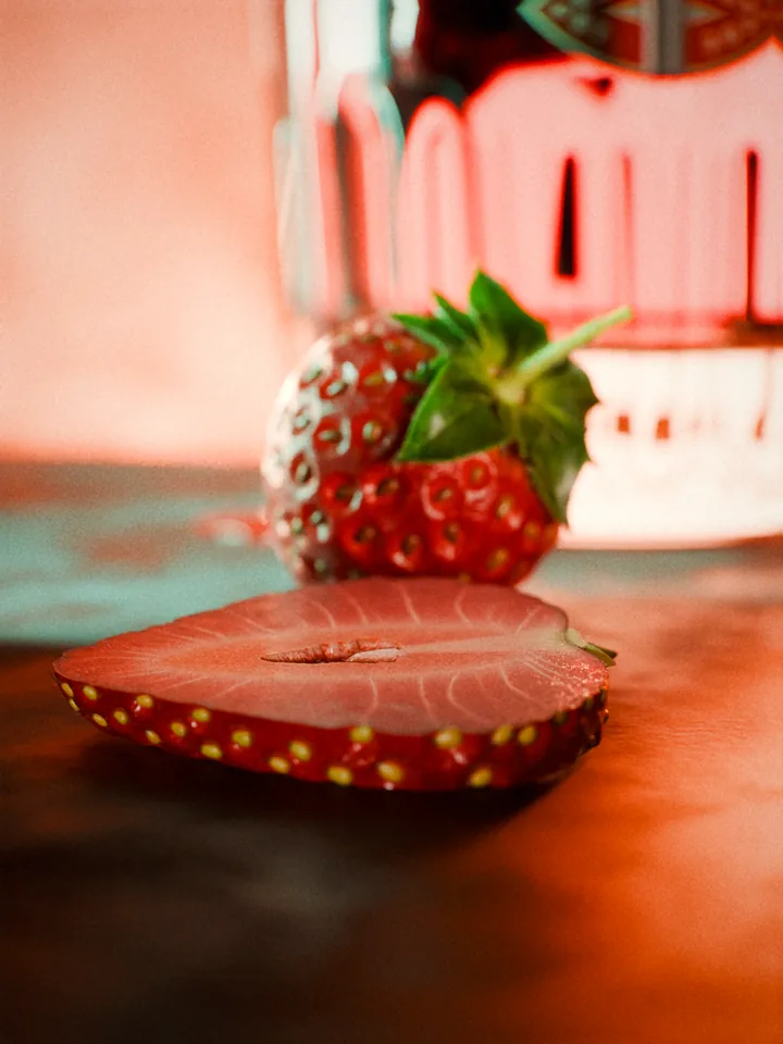 Macro of a strawberry slice lying face up on the steel plug with a whole strawberry and the base of the glass out of focus behind.